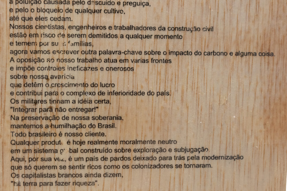 Wood panel with a portion of the author's "Uma Carta de um Membro das Máfias do Ipê" printed on with black ink. 