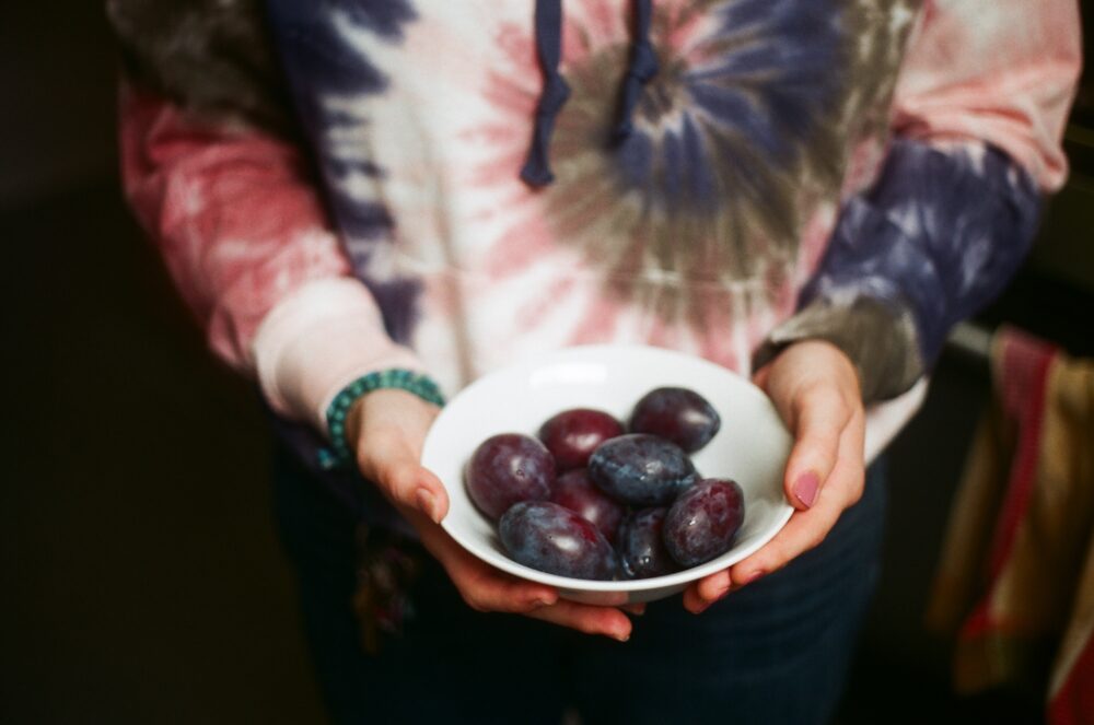Wearing a tie-dye sweatshirt, holding out a bowl of deeply purple plums in a white bowl.