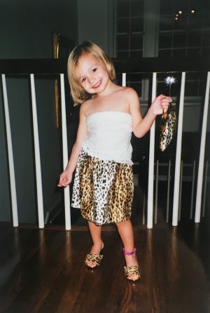 A young girl wearing a leopard print skirt, holding a leopard-print bag, poses for the camera. 
