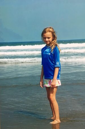 A girl stands on the shore in a swim-shirt, looking down. 