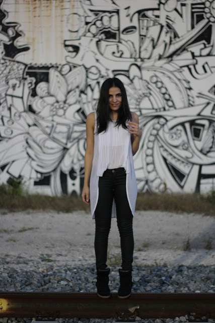 A woman wearing a light top and cardigan and dark jeans standing against a light wall with an abstract mural in thick black paint.