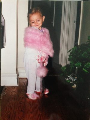 A young girl posing for the camera, wearing a fluffy pink shoulder wrap, pink shoes, and carrying a fluffy pink bag. 