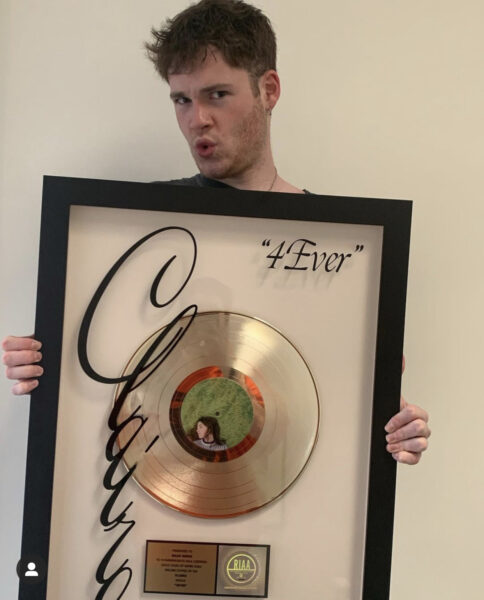 Iz faces the camera displaying his RIAA Certification for his credit on Clairo’s gold single “4Ever.”