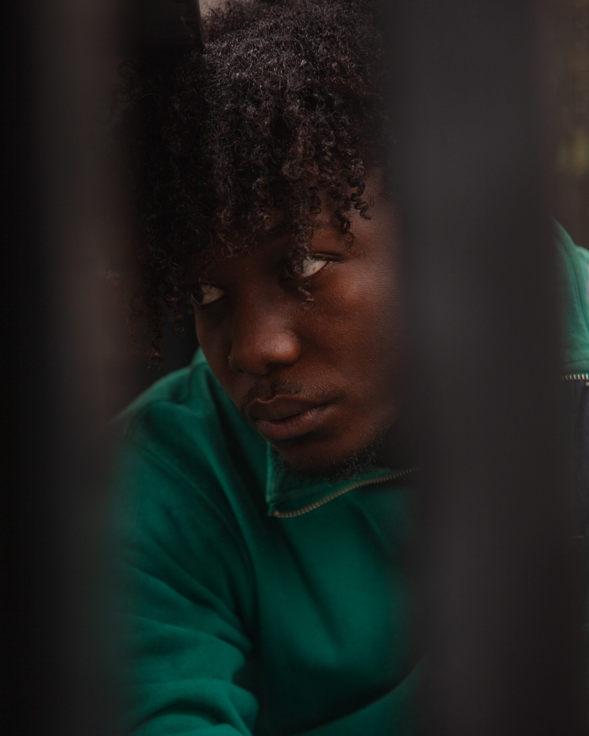 The image is a portrait of a black man with curly hair, wearing a green jacket. The man is looking to the side and not directly at the camera. The photo features a blurred foreground of grey bars, with the focus on the man's face, and a soft, indistinct background