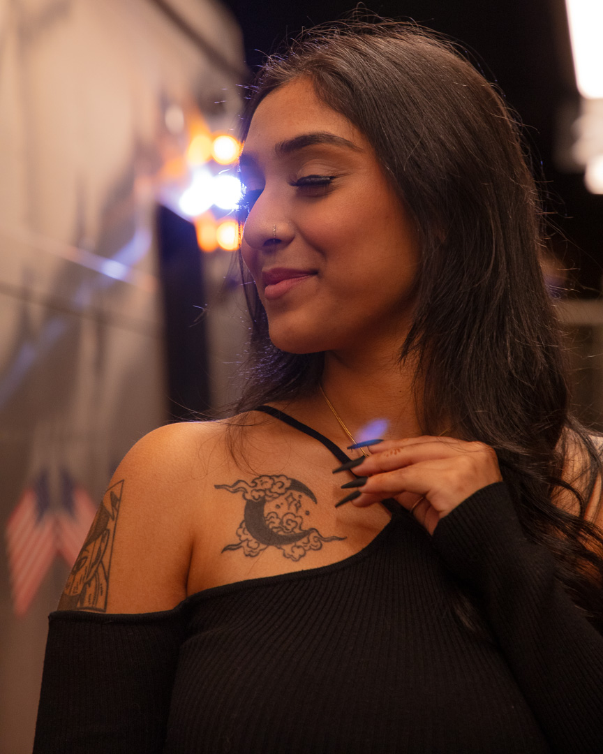 The photograph captures a woman with a nose ring on her left nostril, a serene smile, and her gaze directed downward. She has a tattoo of the moon on her collar/chest area and is wearing a long-sleeved black halter top. Her hand is gently touching a necklace's pendant, drawing attention to the moon tattoo on her collar. There is another tattoo picking over her arm. In the background, the lights of a train create a bokeh effect, contributing to the night-time urban atmosphere of the scene