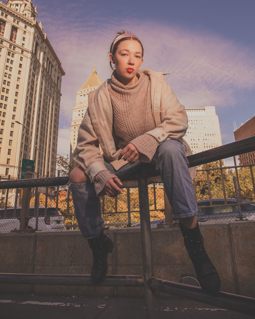 The woman is perched on a metal railing and dressed in a large beige sweater, cuffed denim jeans, and black boots with spike embellishments. She has her hair tied back, adorned with a headband, and her red lipstick is a distinct feature. Her face is turned to the side, yet her eyes are gazing directly at the camera, creating a direct and engaging connection with the viewer. Behind her, the urban backdrop of tall buildings under a softly hued sky in a city environment.
