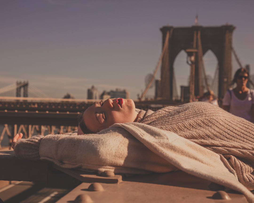 The photo captures a woman lying on her back with her eyes closed, resting on a hard surface, draped with a beige textured sweater. She is wearing red lipstick. In the background, the Brooklyn Bridge spans across the frame under a clear blue sky, with people walking by.
