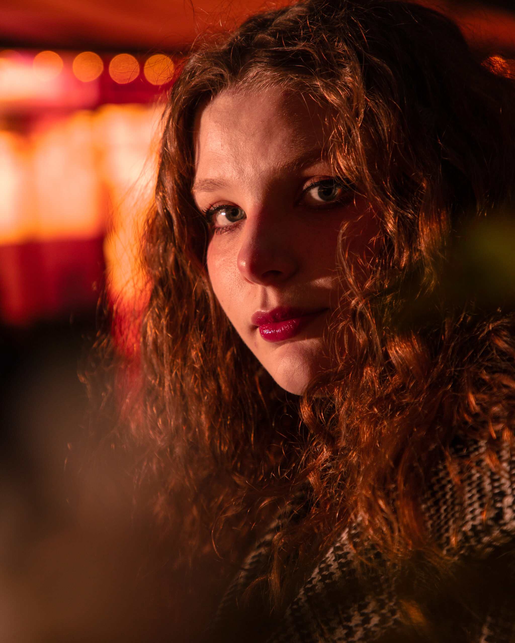 The photo shows a close-up of a woman with curly hair, looking directly at the camera. She has pinkish-red lipstick on, and the lighting casts a warm glow on her face, accentuating her features. In the background, out-of-focus lights create a bokeh effect, contributing to a warm ambiance. The woman is wearing a patterned garment, although it's not fully visible. The overall tone of the photo is warm, with a focus on the woman's face and expression.