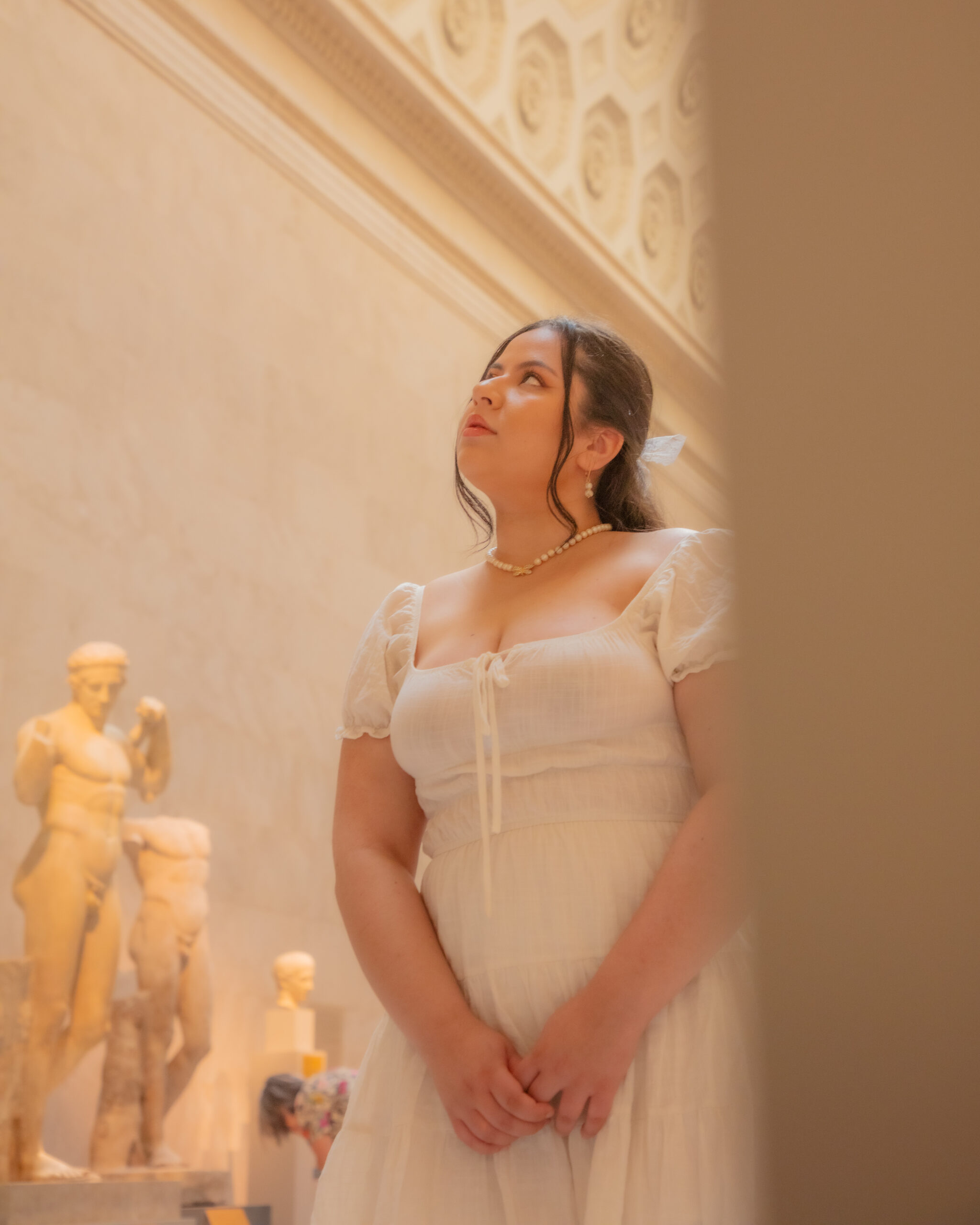 The photograph displays a woman inside a museum, dressed in a white dress with puffed sleeves and adorned with a pearl necklace. She stands with her hands clasped in front of her, looking up thoughtfully. Statues from classical antiquity are visible in the softly lit background, contributing to the cultured ambiance of the setting.