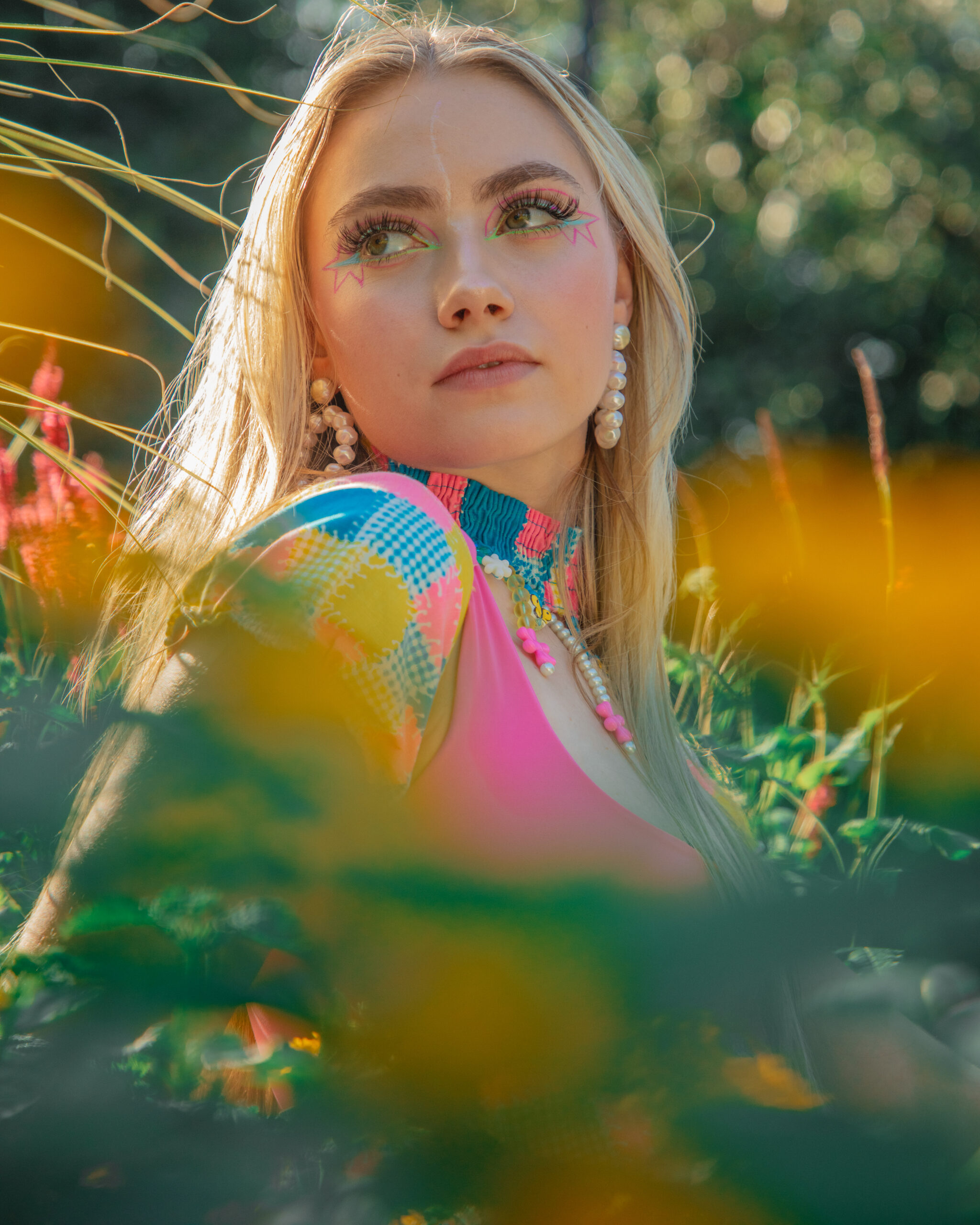 The image features a woman with blonde hair and makeup that includes prominent pink and green details around her eyes. She is wearing large pearl earrings and a colorful garment with pink, blue, and yellow patterns. The woman is gazing upwards, and the photo is taken from a lower angle with some yellow flowers in the foreground, which are blurred, while the woman is in focus. The background is a sunlit environment
