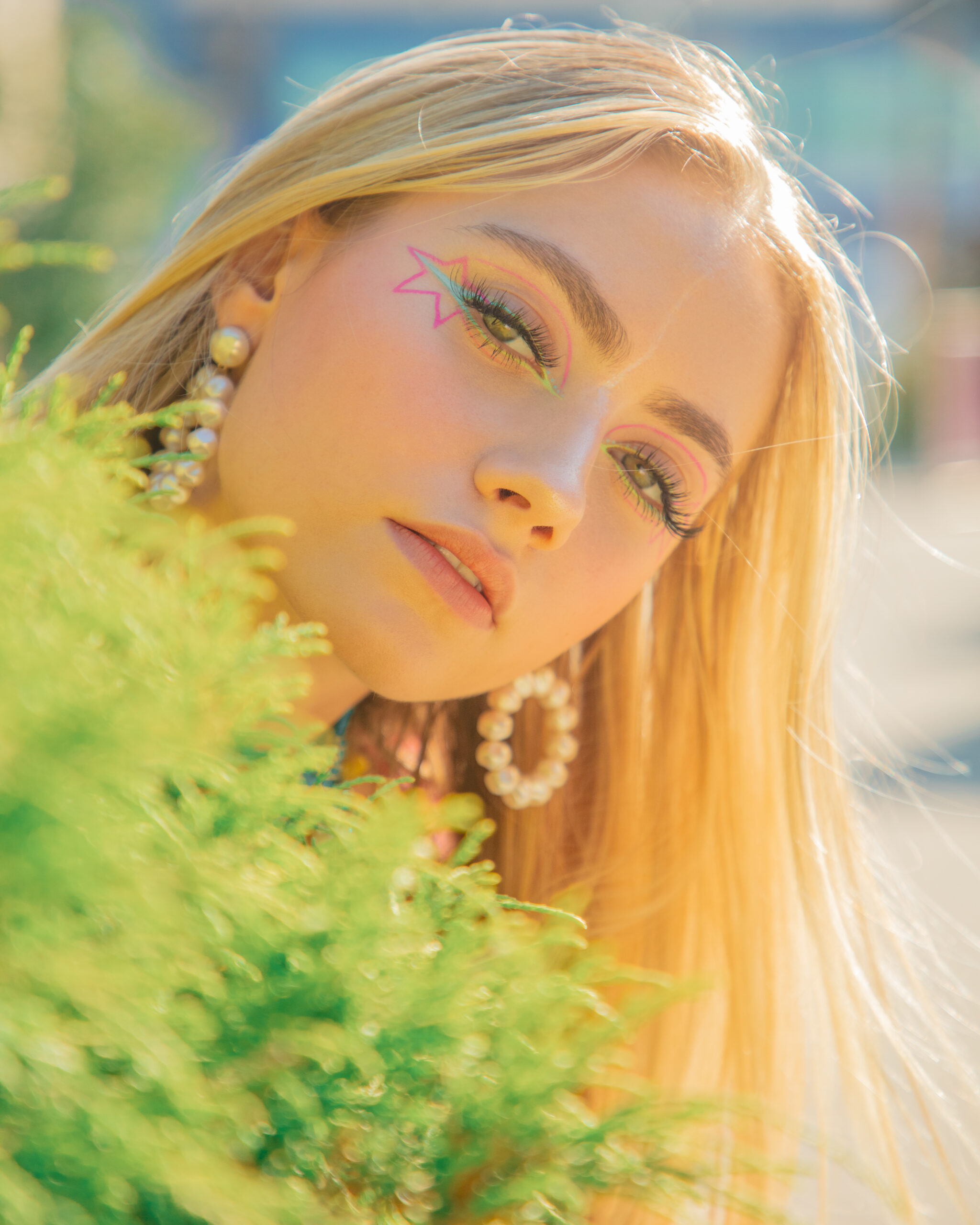 The image is a close-up portrait of a woman with blonde hair and creative makeup featuring a pink star near her right eye. She is wearing large pearl hoop earrings and there's a soft focus on greenery in the foreground. The background is overexposed, giving the image a bright, airy feel. The woman is looking slightly to the side, and her facial expression is serene. The lighting on her face is soft and warm