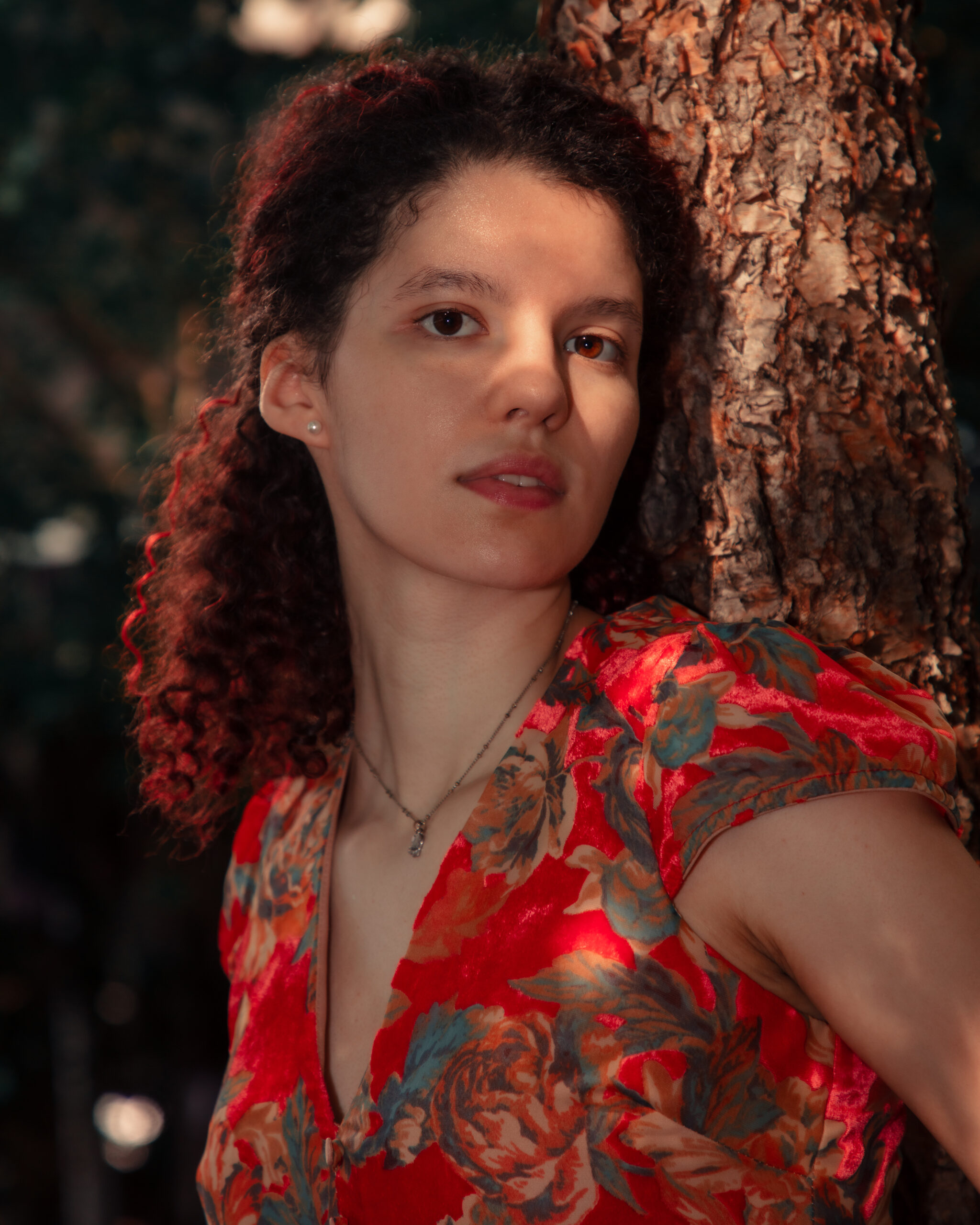 The image is a portrait of a woman with shoulder-length brown curly hair leaning against a tree. She has a neutral expression and is looking slightly away from the camera. She is wearing a red floral top with a V-neckline and a delicate necklace. The lighting is warm, casting soft shadows on her face, and the tree bark provides a textured backdrop. One of her eyes is in shadow, the other is brightly lit up by the sun, making it seem as if she has heterochromia. The overall tone of the image is warm with a focus on natural colors