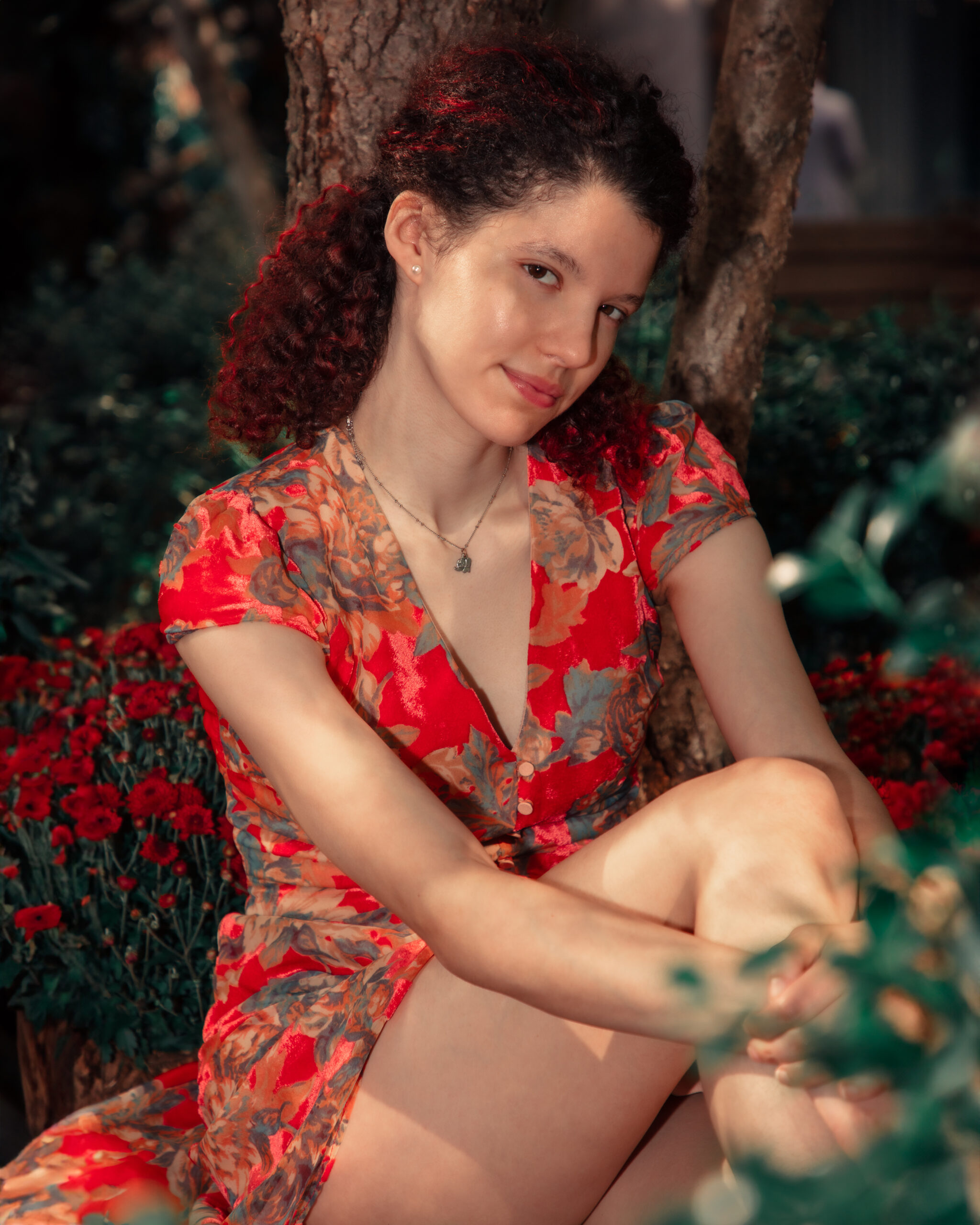 The image is a portrait of a woman sitting with one knee drawn up, leaning against a tree. She has curly brown hair  with red highlights, is smiling slightly, and is looking at the camera. She's wearing a red dress with a floral pattern and a V-neckline. She is wearing a delicate necklace. The environment suggests an outdoor setting with foliage and red flowers in the background. The lighting is warm, enhancing the red and green tones in the scene.