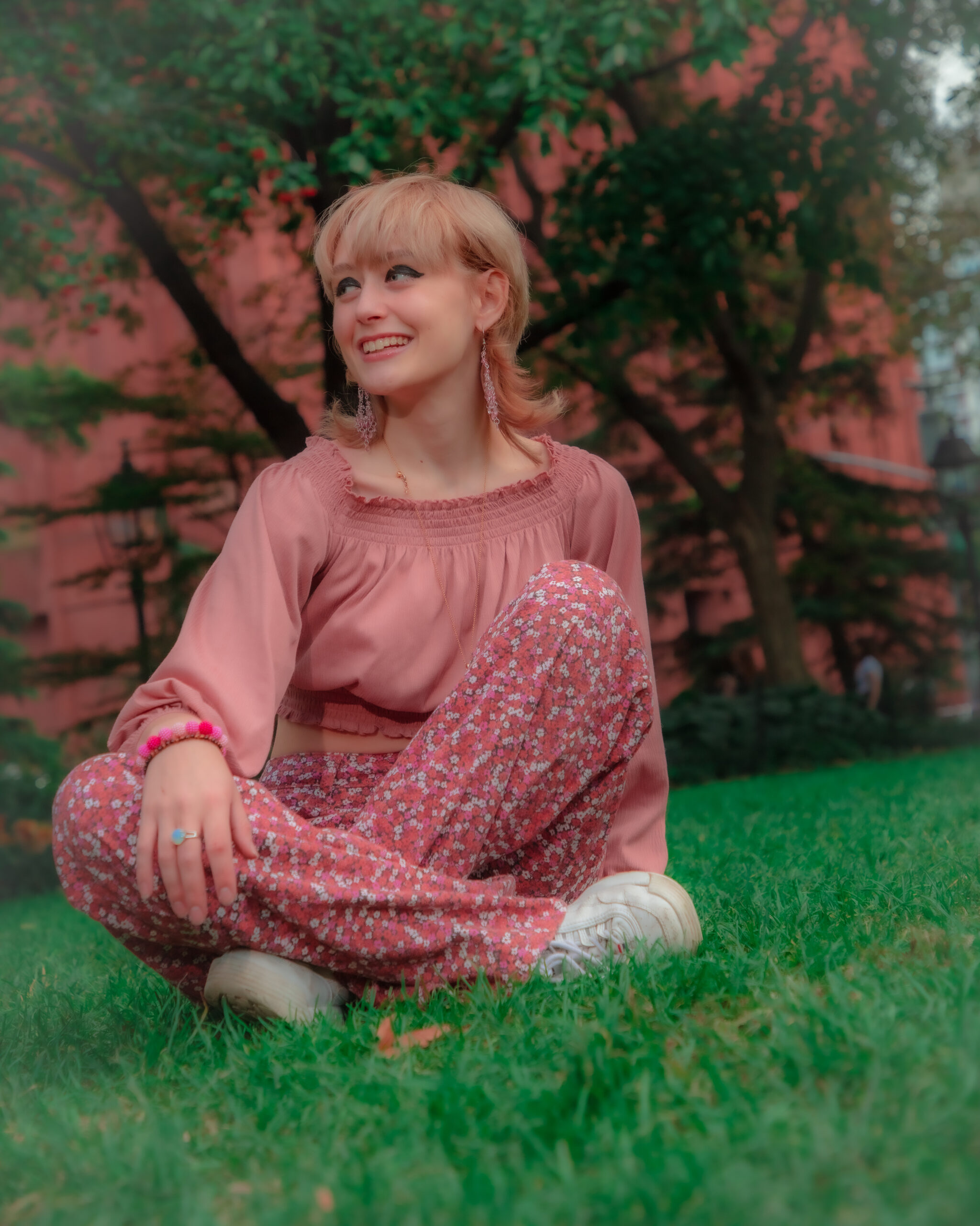 The image displays a woman sitting cross-legged on grass, with a smiling expression and looking upwards to her right. She has blonde hair with bangs, and she's wearing a pink blouse paired with floral-patterned trousers. She has on white sneakers and is adorned with earrings and a ring. The background features green foliage and a reddish-pink building, which together with the woman's attire and the green grass, creates a colorful and relaxed outdoor setting