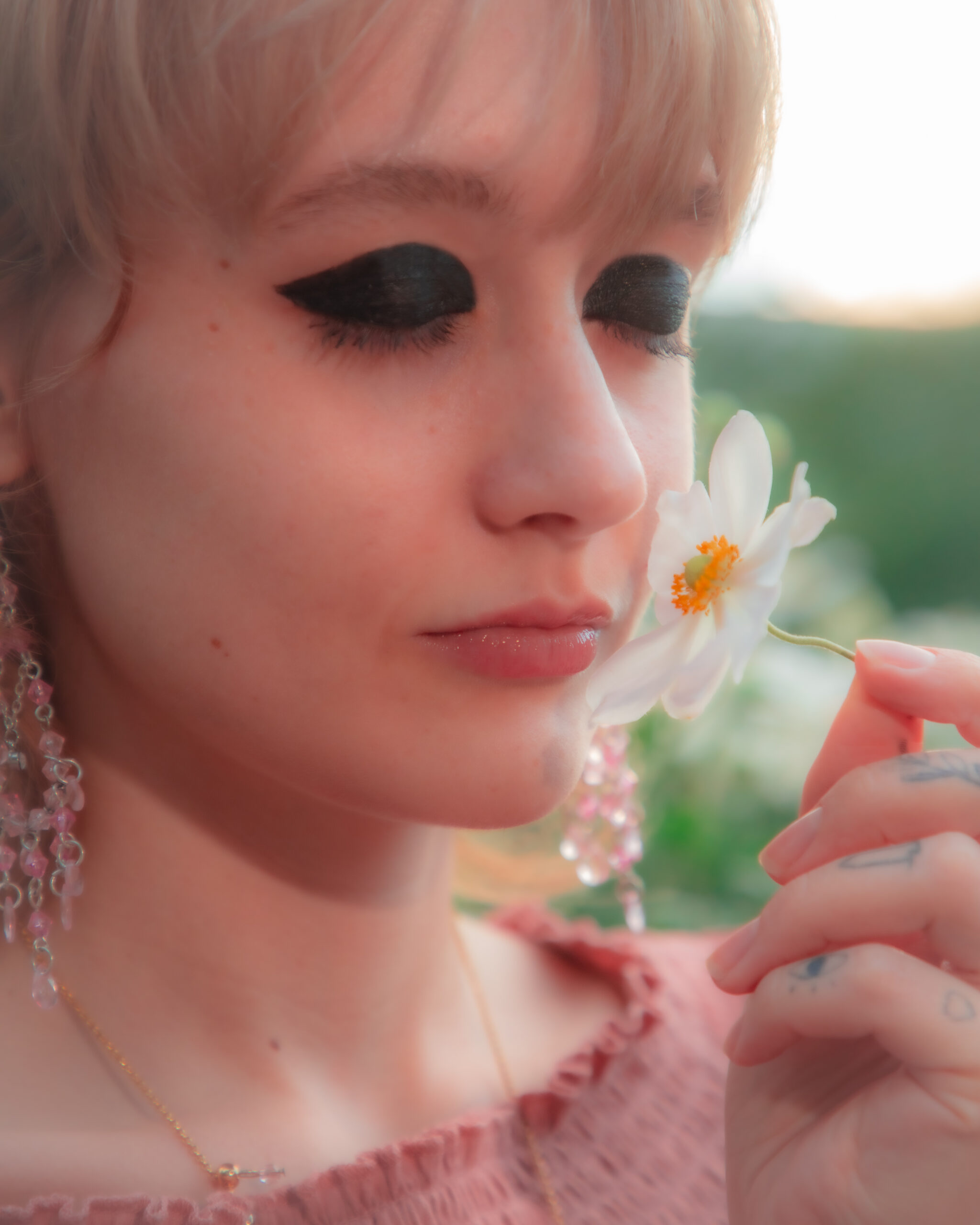 The image is a close-up of a woman holding a white flower close to her face, smelling it with closed eyes. She has prominent black eye makeup, blonde hair, and is wearing a pink blouse. Her earrings are long and beaded, and a delicate necklace is visible. The background is softly blurred with a warm, glowing light, suggesting a setting with soft natural lighting, possibly during golden hour