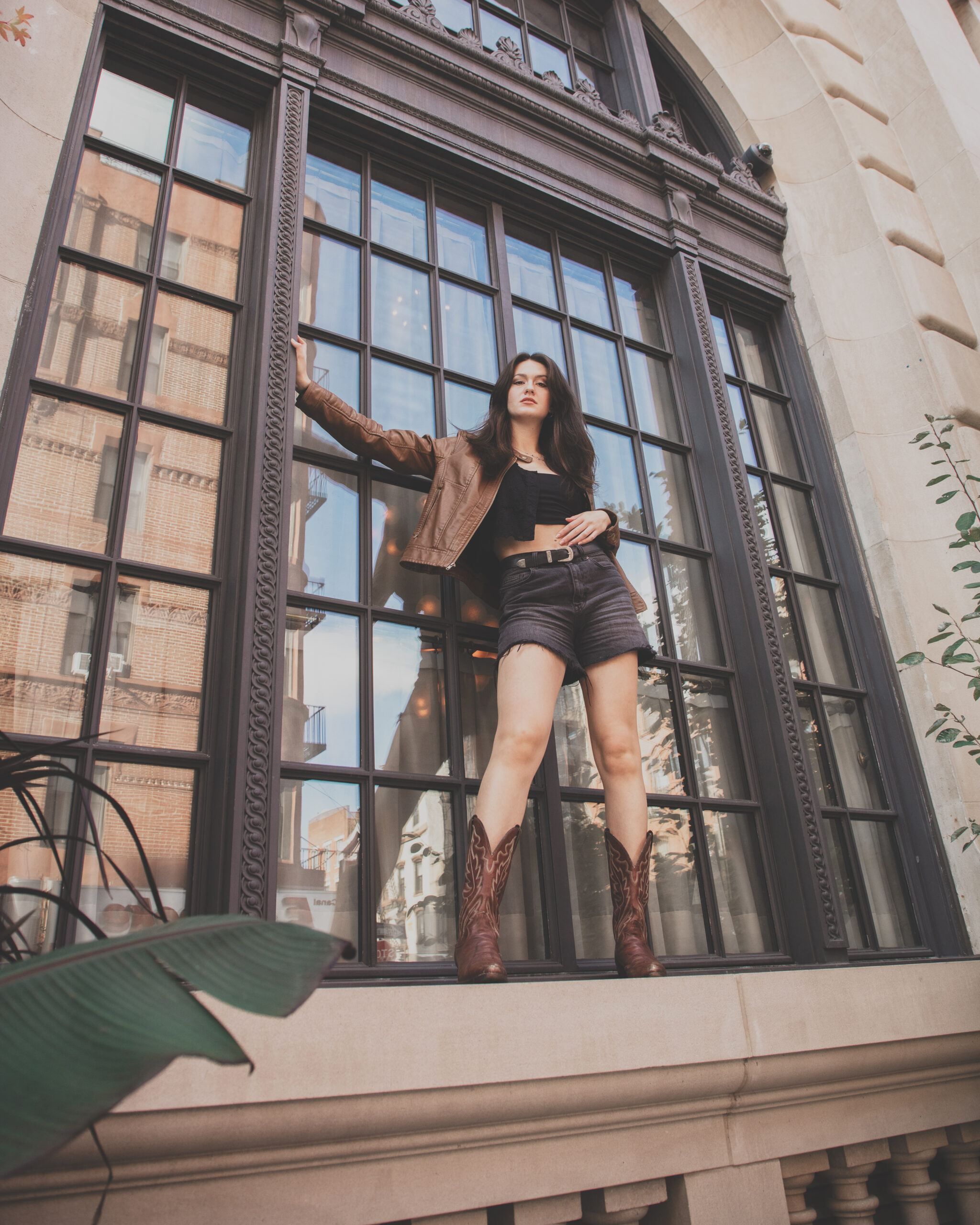 The image depicts a woman standing in a very large window frame with her left hand extended to the side of the frame. She has dark hair and is wearing a brown jacket, black top, dark shorts, and brown cowboy boots. The window reflects the street and buildings opposite. There is a large leaf in the foreground on the left side of the frame, adding a touch of greenery to the urban setting