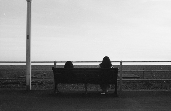 Two people sitting on a bench looking out on a dock, photographed from behind in black and white. 
