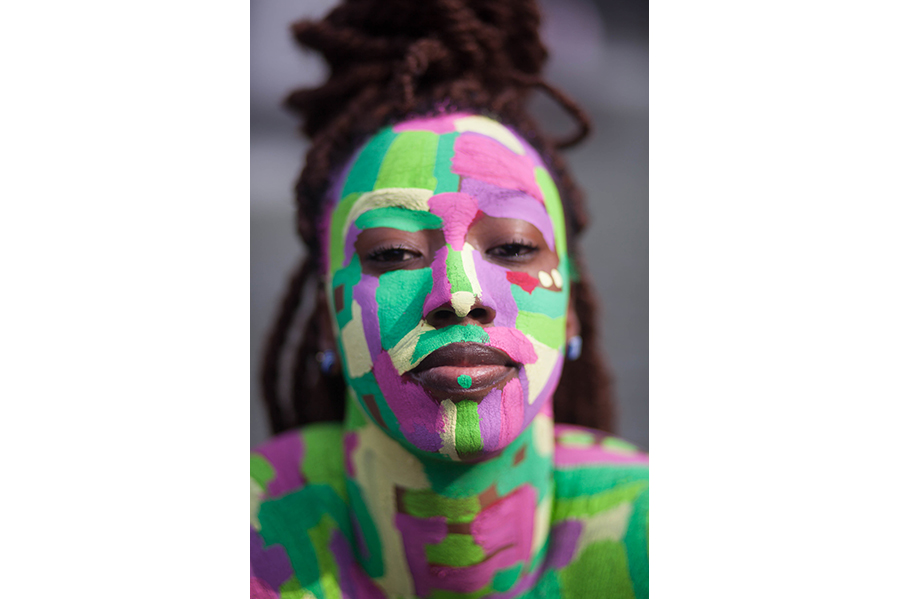 A women with locs and her face painted with green, magenta and yellow swatches gazes into the camera. 