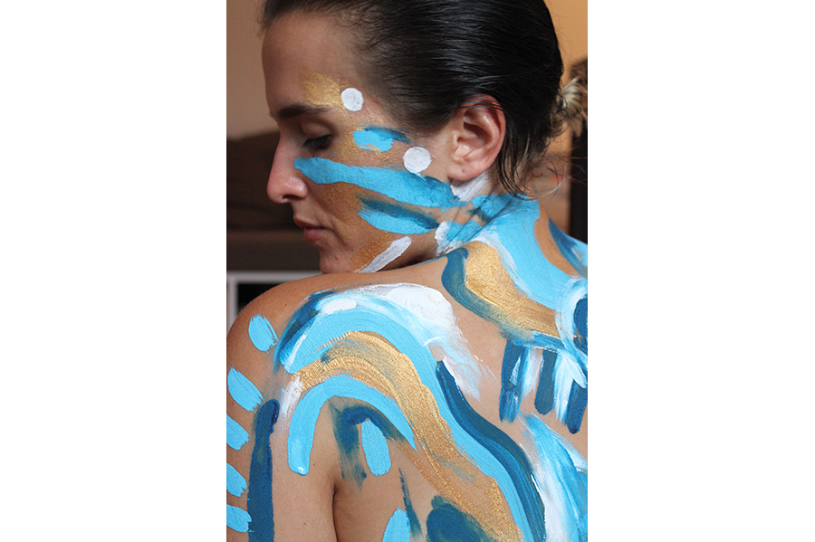 A close-up of a woman looking over her shoulder, her back and face painted with light blue, dark blue, and white. 