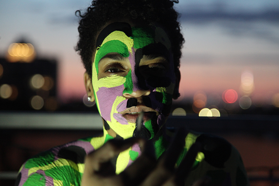A young person with green, light green, and purple paint on their face and shoulders reaches toward the camera at sunset. 