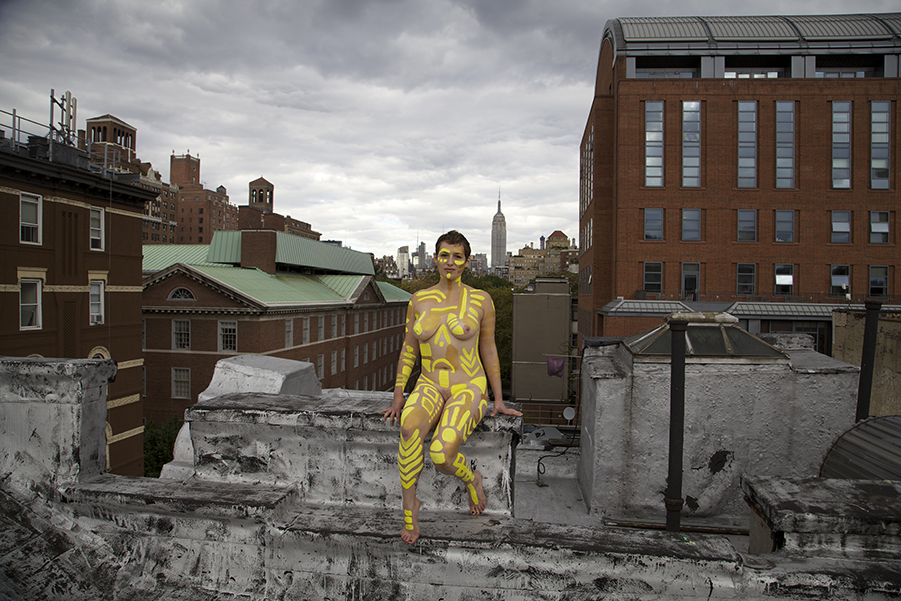 A woman sits on the ledge of an unfinished building, her body panted with bold yellow lines of paint.