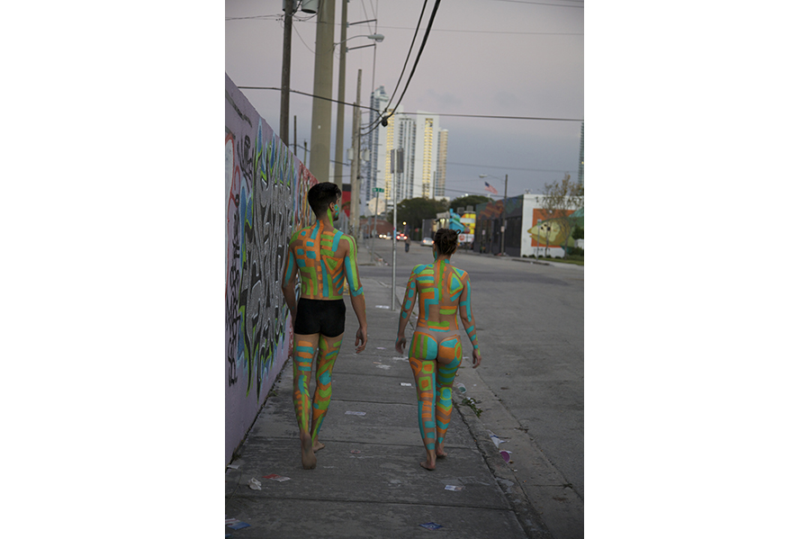A man and woman with green, blue and orange body paint walk down the street.