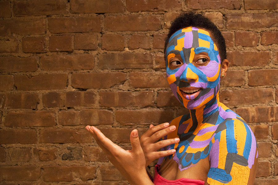A person with blue, green and yellow body paint sits in the center of a stone street and gazes into the camera. 