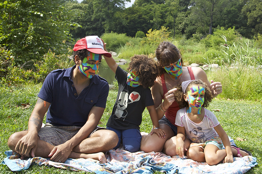 A family sits on a blanket, with blue, green, orange, and brown face paint.