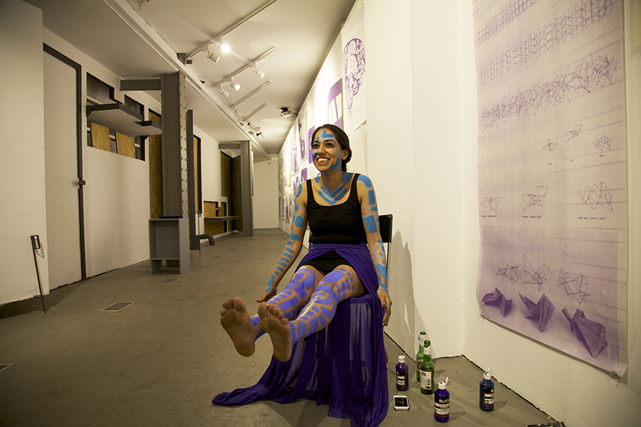 A woman wearing a leotard, purple maxi skirt, and blue and purple body paint sits on a chair. 