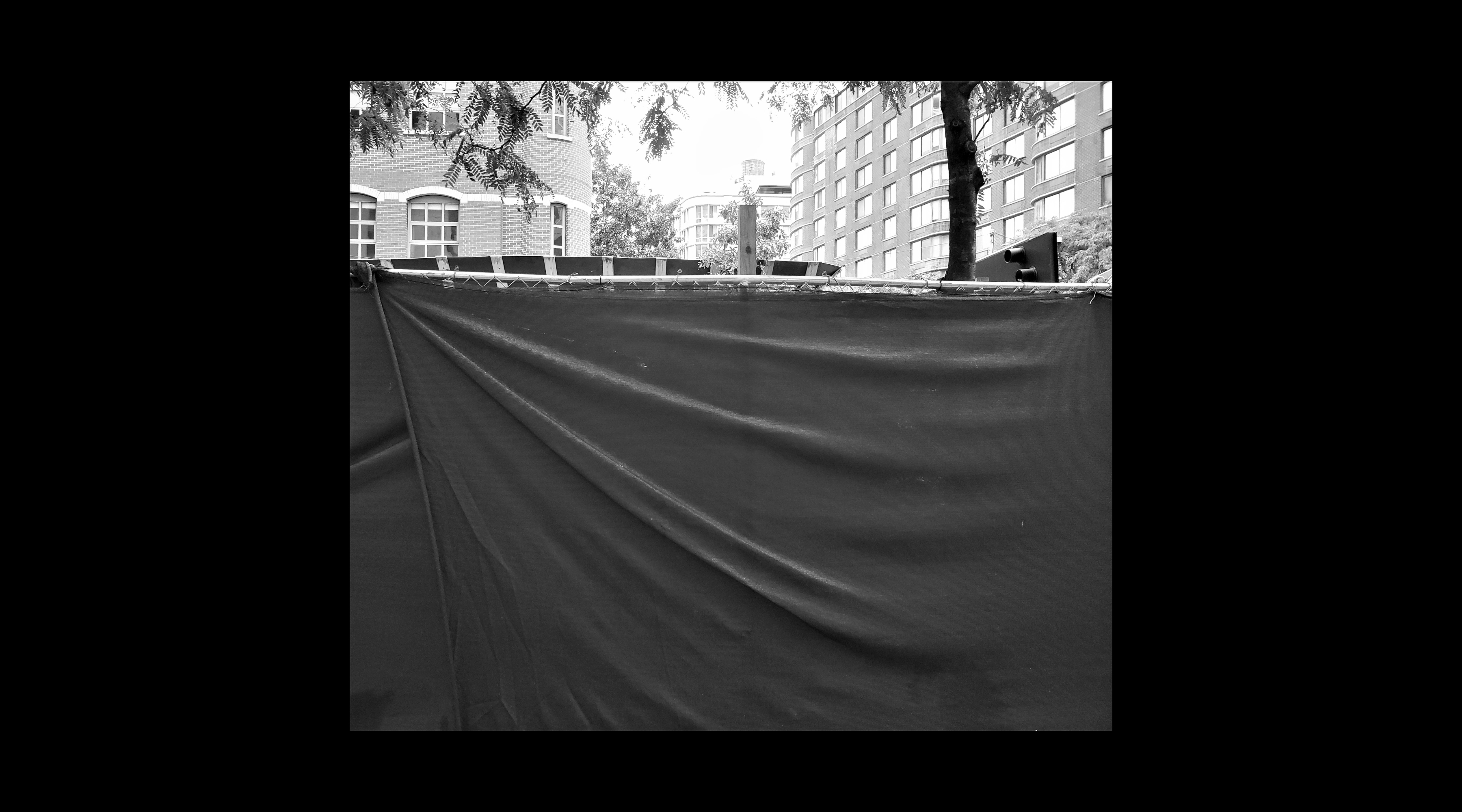 A chainlink fence, covered by a tarp; above it in the top third of the frame, two trees and buildings (in black and white)