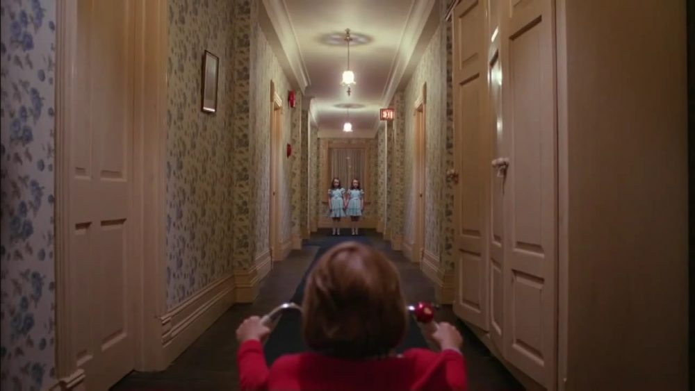 A young boy on a tricycle encounters young twin girls at the end of a narrow hotel hallway.