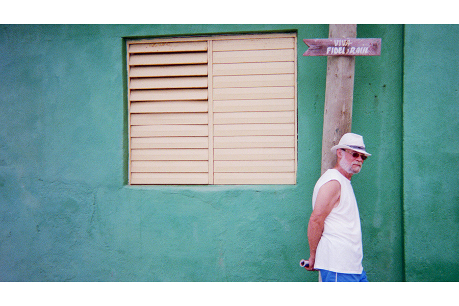 A man in a white tank top, sunglasses, and white hat stands against a blue-green wall, gazing toward the camera. 