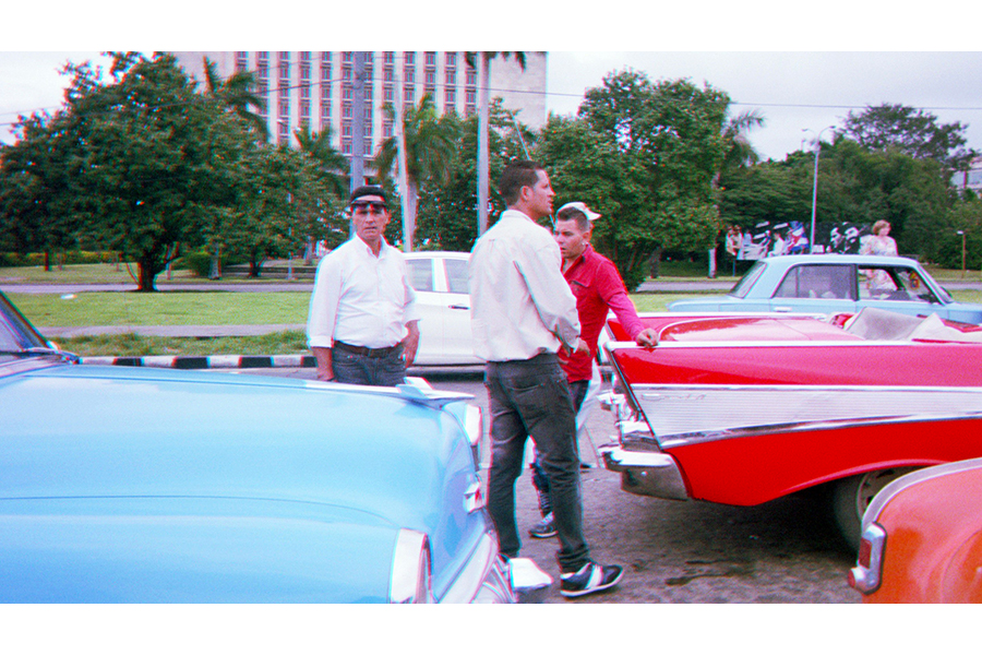 Three men stand between two cars, both with candy paint (one light blue and the other bright red).