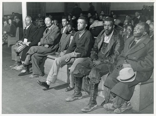 Photograph of the first row of spectators in a court room, one man meets the camera with his gaze.