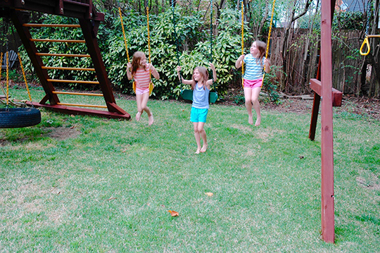 Color photograph of young girls on outdoor swing set, grass and plants in background