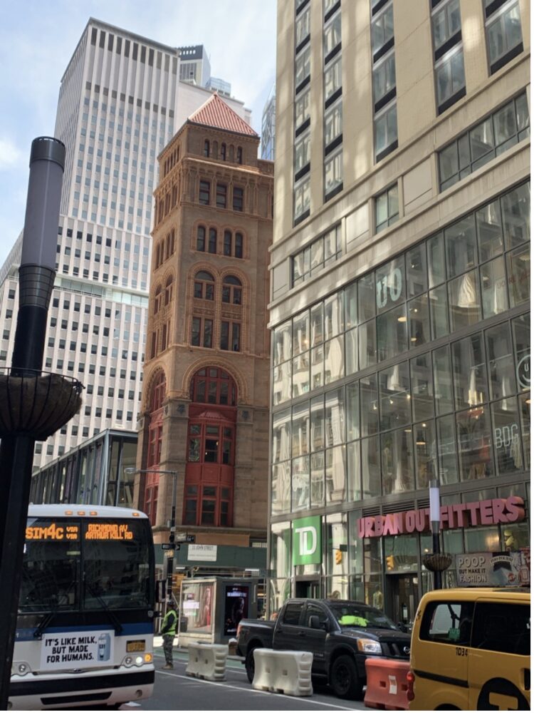 View up Broadway where each building in sight is from a different era, in a different architectural style. The foreground of the image depicts traffic and construction.