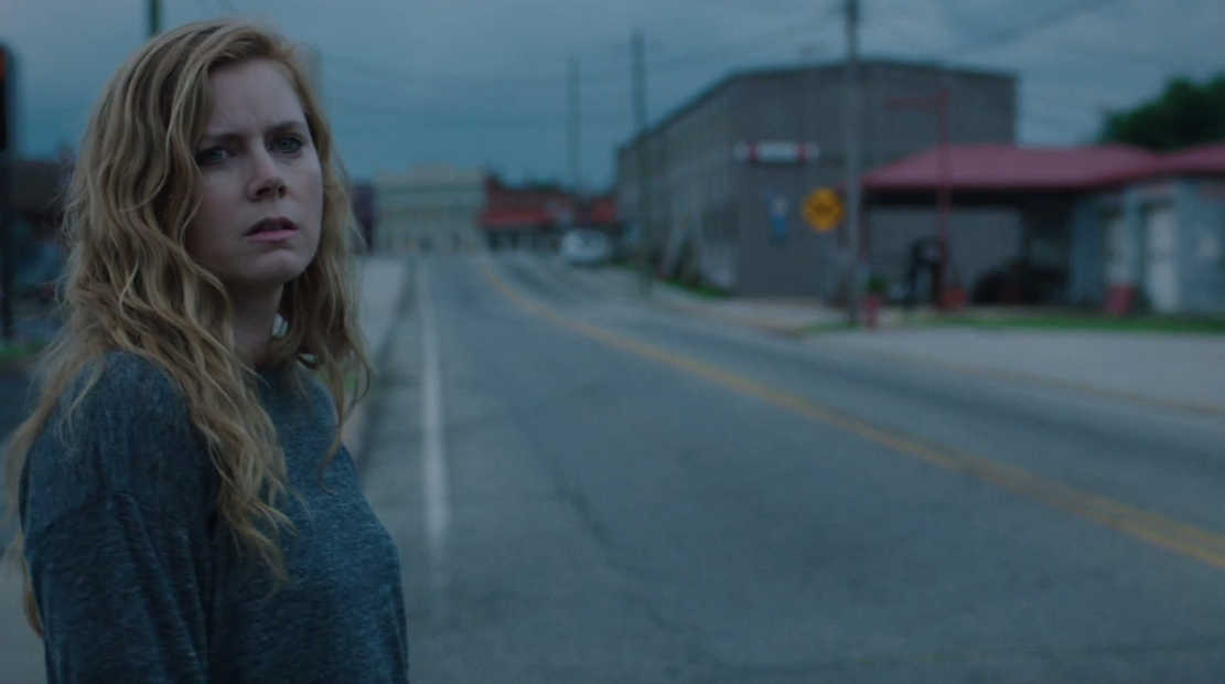 Amy Adams as Camille Preaker stands at the side of the road in the foreground; in the background, the road, and some low brown buildings appear in soft focus against a gray sky