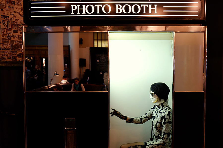A woman with white bandages around her head and white gloves sits in a photo booth. 