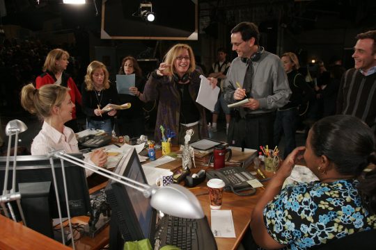 photograph of Julie Larson, center, speaking with two actors seated at desks on a TV set; crew in the background