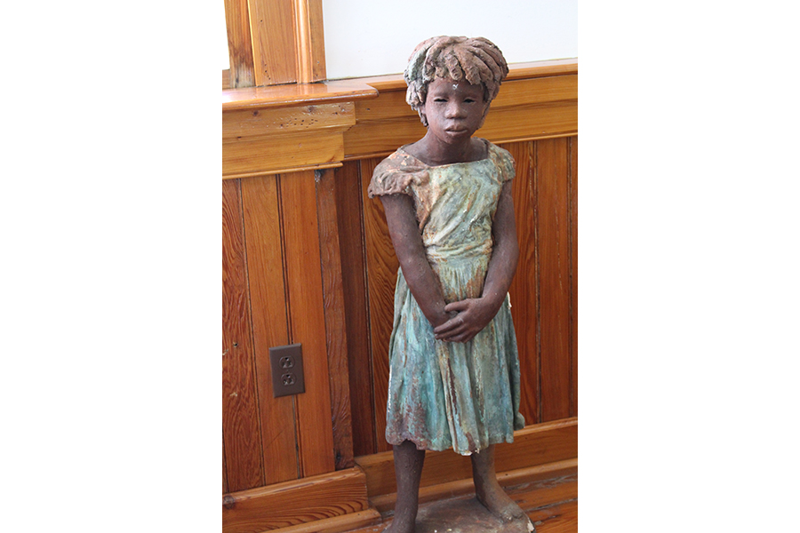 Artist Woodrow Nash has captured one of several Children of the Whitney in a clay sculpture. The little girl poses beside a church window, hands folded in front of her, greeting visitors with grave, cavernous eyes . The sole touch of color can be found in her simple turquoise dress, streaked with mud. 