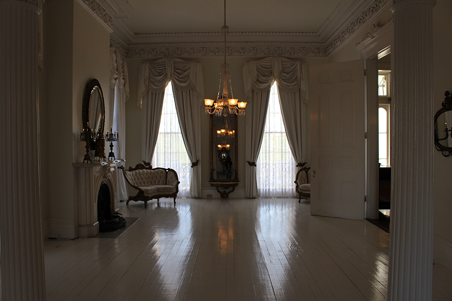 Living up to its namesake, the White Ballroom boasts white molding reminiscent of a garland of English roses, white drapes with a hint of fringe, white wooden floors, a white fireplace, and a matching set of white loveseats framed in dark wood. Though light in color, the room is rather dim—sunlight is filtered through sheer curtains pulled over a pair of windows, two small candelabras emit only minimal light from their place atop the fireplace, and the warm glow of the crystal electric chandeliers fails to illuminate more than a tiny bit of space. 