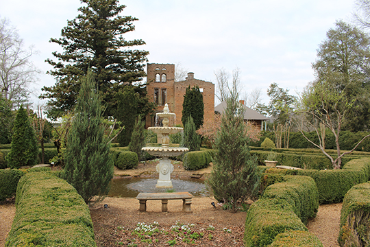 The gutted brick facade of a grand building stands in a landscaped garden behind a fountain, hedges, and trees.