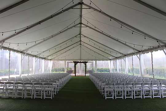 Interior of a large wedding tent empty, fully covered, with clear plastic sides, row seating of white chairs, globe lights, and a simple wooden canopy at the altar