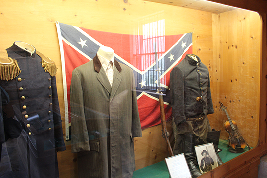 In a glass display case, Confederate and Union army uniforms are displayed on mannequin forms before a confederate flag. A rifle, a fiddle, boots, and a black-and-white portrait of a solider stand to the side. 
