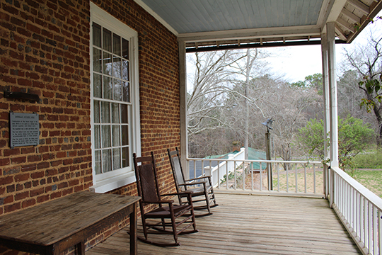 Two rocking chairs, empty, on a wooden, covered porch, the red-brick building facade in the background