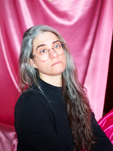 A woman with long gray hair and wire-rim glasses wearing a solid black shirt and a contemplative expression against a bright pink curtain.