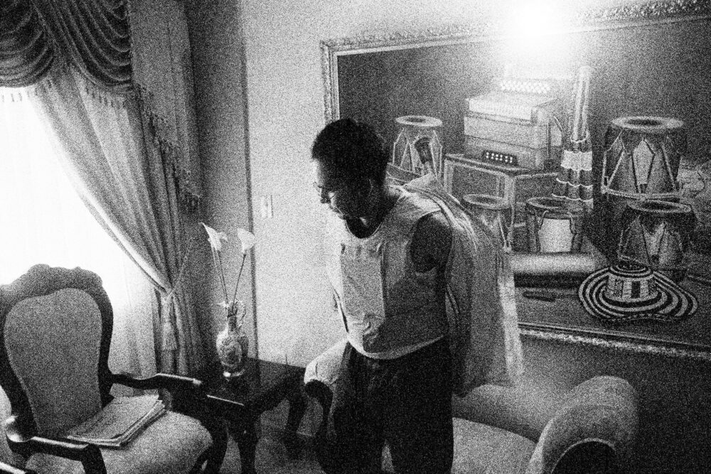 A man takes off his shirt, revealing a bulletproof vest in a lavishly decorated room.