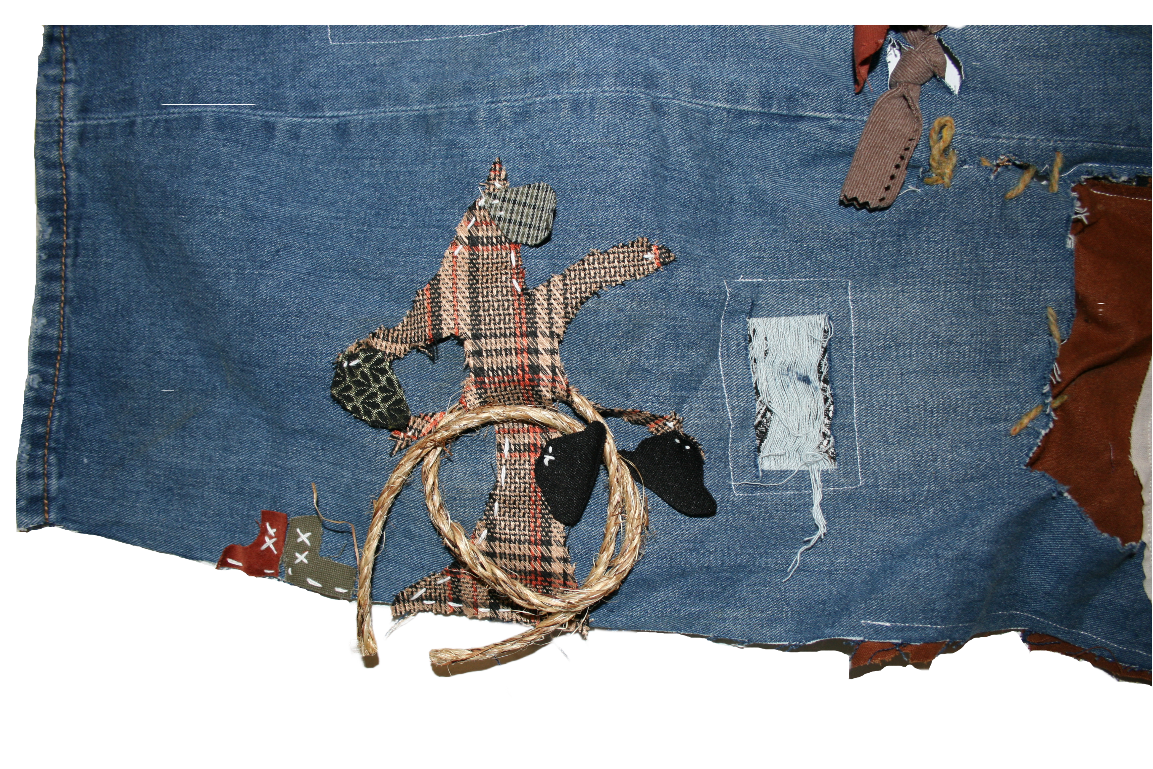 Close up photo of the bottom left corner of the piece. There is a tree made of brown patterned fabric sewn onto the denim. The tree has two black hats, two green leaves, and a brown rope hanging from it. There is a pair of mismatched boots sewn to the left side of the tree and a rectangle of distressed denim to the right side.
