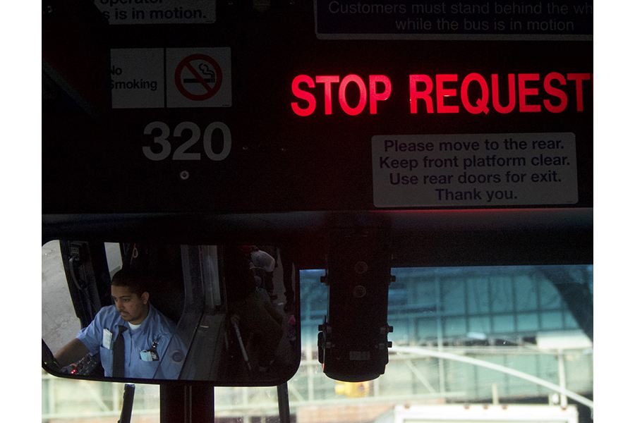 Bronx bus driver, in a blue uniform, seen through the bus’s rearview mirror. 
