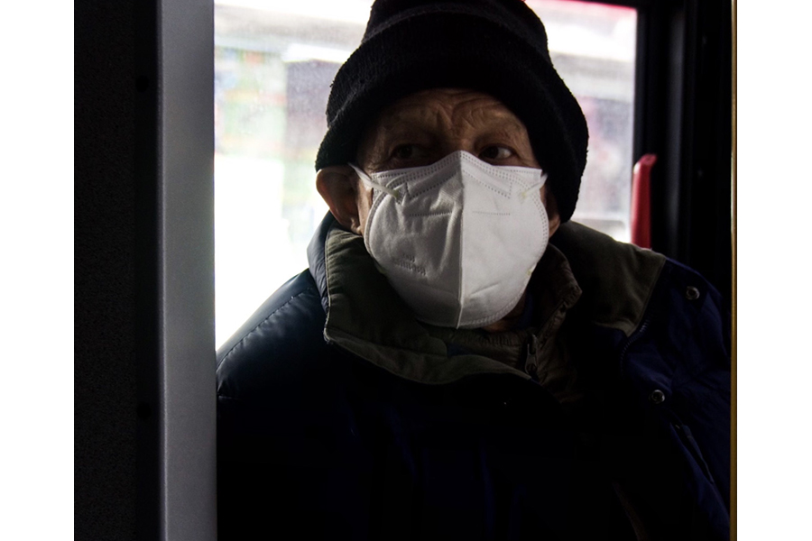 Portrait of an elderly man on the bus wearing a blue heavy jacket with half his face hidden behind a face covering, looking out of the bus into the Manhattan streets.